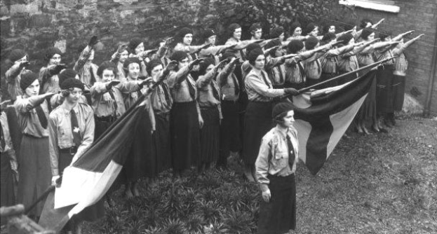 Schwarz-weiß-Fotografie einer Gruppe von Frauen der Frauenwahlrechtsbewegung, die zusammenstehen und Fahnen halten, mit Bäumen im Hintergrund.