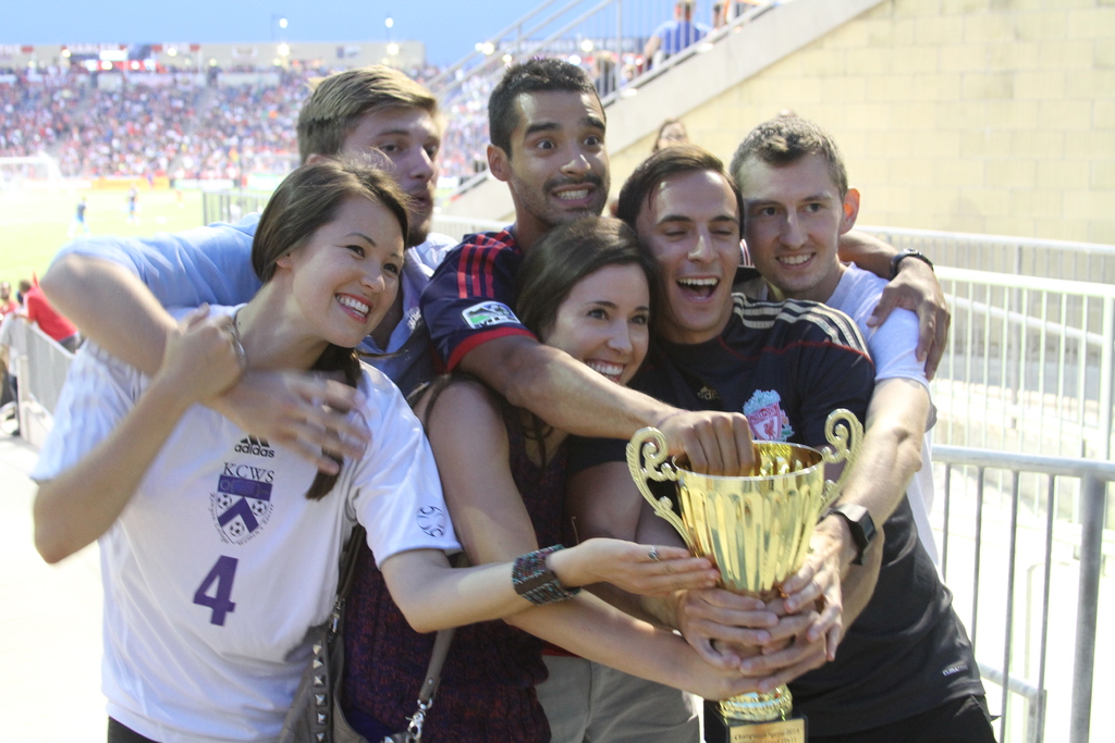 Sechs Personen, darunter zwei Frauen und vier Männer, die einen Pokal halten und lächeln, posieren für ein Foto vor einem Eisengeländer, mit einer Wand und einem Geländer oben rechts, Menschen und Lichter im Hintergrund, möglicherweise in einem Stadion.