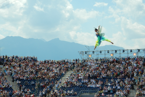 Eine Frau in einem grünen Kleid fliegt über eine sitzende Menge in einem Stadion, mit Bergen, Wolken und einem klaren blauen Himmel im Hintergrund und einer beleuchteten Bühne auf der rechten Seite.