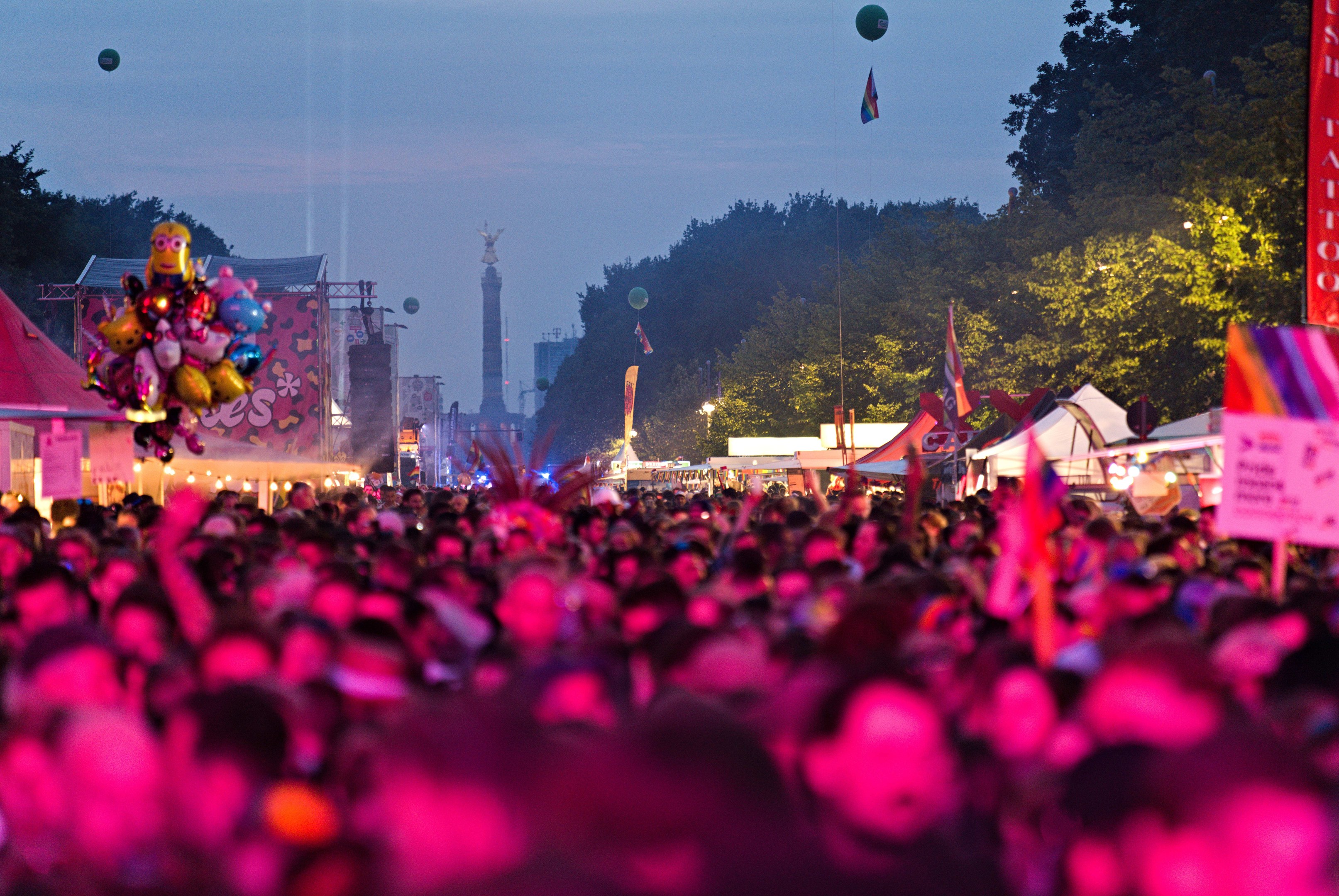 Eine große Menge bei einem nächtlichen Musikfestival mit bunten Lichtern, Ballons, Zelten, Schildern, Fahnen, Bäumen und einem sternenklaren Himmel im Hintergrund.
