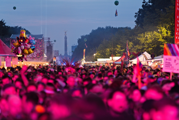 Eine große Menge bei einem nächtlichen Musikfestival mit bunten Lichtern, Ballons, Zelten, Schildern, Fahnen, Bäumen und einem sternenklaren Himmel im Hintergrund.