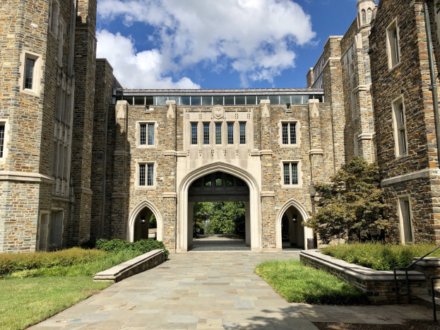 Großes steinernes Universitätsgebäude mit einem Weg, umgeben von grünem Gras, Pflanzen, Bäumen und einem bewölkten Himmel.