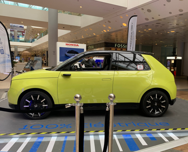 Gelbes wasserstoffbetriebenes Elektroauto vor einem Gebäude mit Absperrpfosten, Seilen, Bannern, einer Person im Auto, einer Säule, stehenden Menschen, Schildern, einer Topfpflanze, Deckenleuchten und Dachventilatoren.