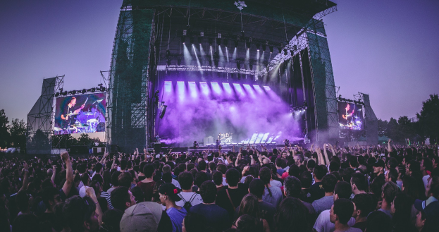 Eine große Menschenmenge bei einem Musikfestival mit einer Bühne, auf der Musiker spielen, beleuchtet von Lichtern und umgeben von Bildschirmen, vor einem Gebäude, Bäumen und einem klaren blauen Himmel.