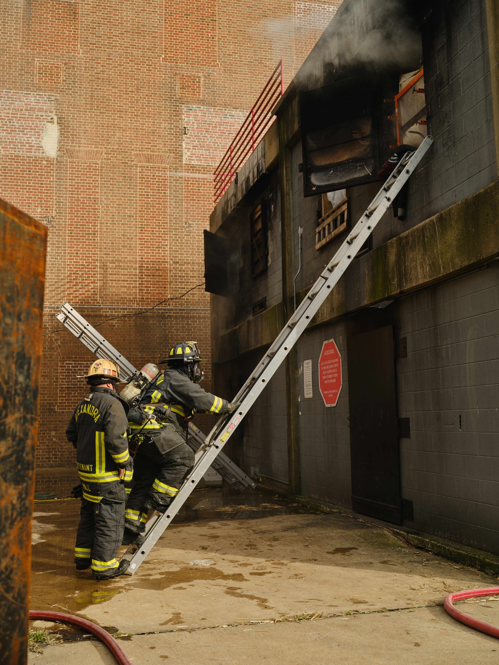Feuerwehrleute in Helmen klettern eine Leiter hinauf zu einem rauchgefüllten Gebäude, mit einem Metallobjekt links und Rohren an der Basis.