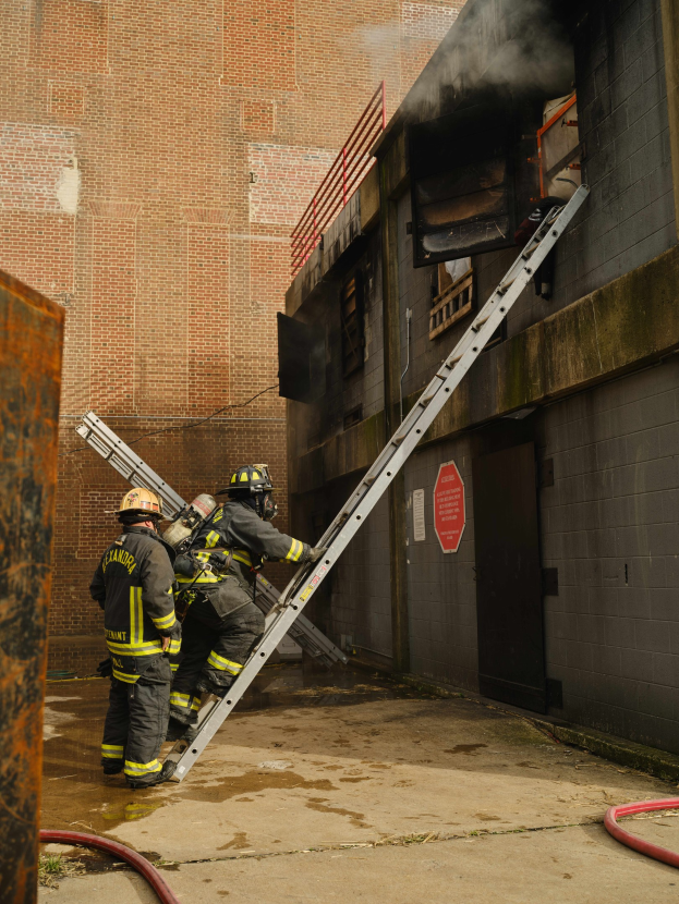Feuerwehrleute in Helmen klettern eine Leiter hinauf zu einem rauchgefüllten Gebäude, mit einem Metallobjekt links und Rohren an der Basis.