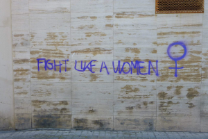 Eine Wand mit den Worten "Kämpfe wie eine Frau" in Blau gesprayt, mit einem Fenster oben und einem Boden unten.