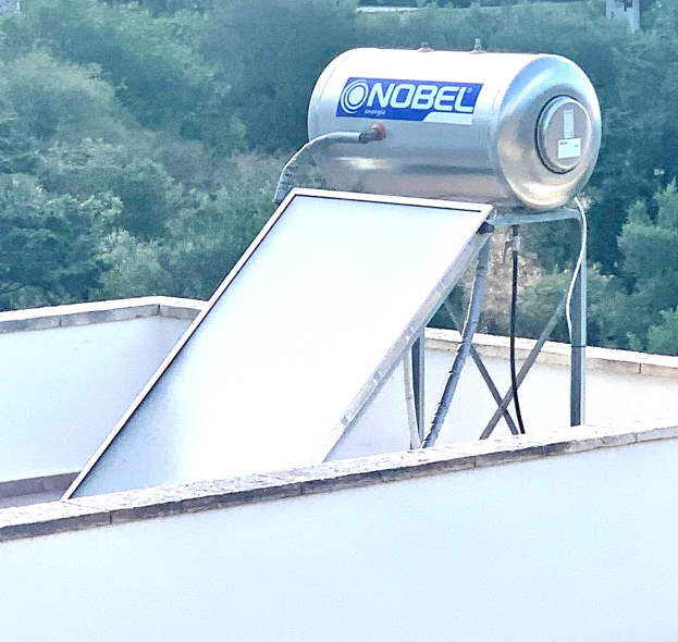 Solarheizung auf dem Dach eines Gebäudes mit einem angebauten Speicher, vor einem Hintergrund aus Bäumen.