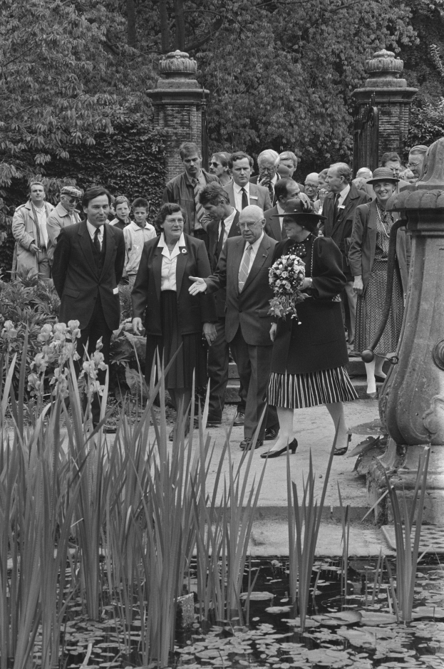 Schwarzes und weißes Bild einer Gruppe von Menschen, die sich um einen Teich in einem Garten versammelt haben, mit einer Frau in der Mitte, die einen Blumenstrauß hält, und einem Mann in einem Anzug rechts von ihr, umgeben von üppigen Pflanzen, Bäumen und Säulen.