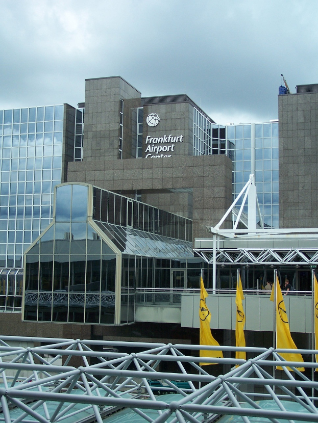 Frankfurt Airport in Frankfurt, Deutschland, ein großes Gebäude mit Glaswänden und Text, umgeben von gelben Flaggen und Eisenstangen, mit einem Himmel voller weißer, flauschiger Wolken im Hintergrund.
