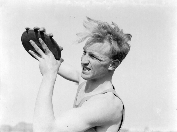 Altes Schwarz-Weiß-Foto eines Mannes in einem weißen Hemd und dunklen Hosen, der einen Fußball hält und einen entschlossenen Ausdruck und zurückgekämmtes Haar hat.