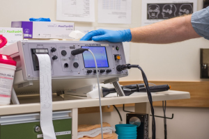 Eine Person in blauen Handschuhen arbeitet an einem medizinischen Gerät an einem unordentlichen Tisch in einer Krankenhausumgebung mit Papieren an der Wand im Hintergrund.