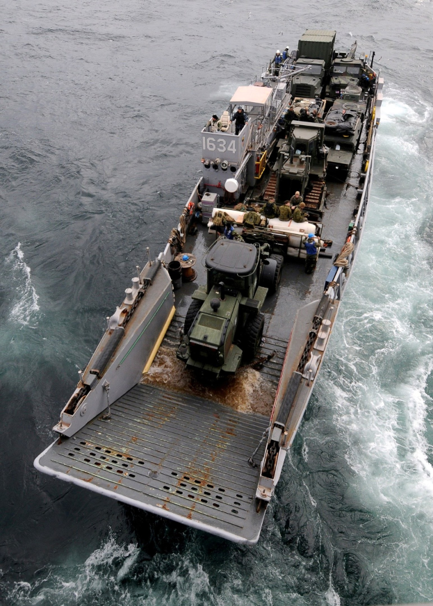 Luftaufnahme eines großen Bootes auf dem Meer mit einem Militärfahrzeug darauf, umgeben von Menschen und Equipment, mit einem Wasserzeichen unten.