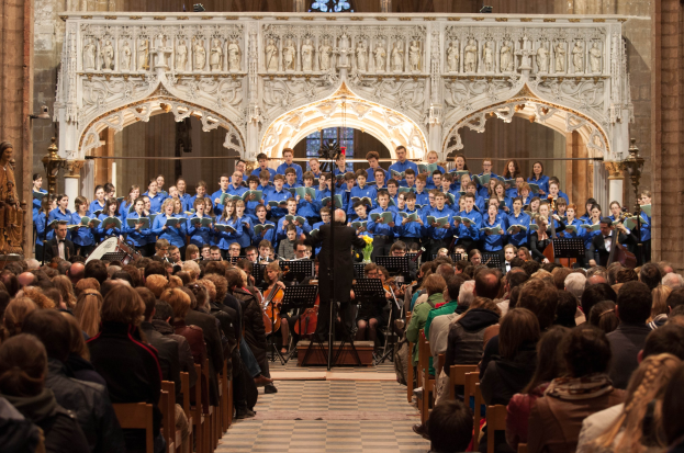 Eine große Gruppe von Menschen steht vor einem sitzenden und stehenden Chor in einer Kirche, mit einer zentralen Person, die ein Mikrofonständer hält. Hinter ihnen befindet sich eine Wand mit Skulpturen und einem Glasfenster, und links sind Statuen zu sehen.