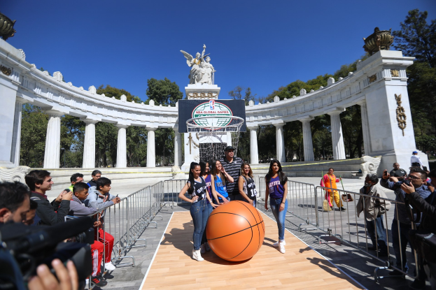 Gruppe von Menschen um einen Basketball auf einem Platz mit einem Netz, Geländern an den Seiten und einem Hintergrund mit Säulen, Statuen, Bäumen und einem klaren blauen Himmel; einige Individuen halten Kameras, was auf eine Teilnahme an den NBA Global Games hinweist.