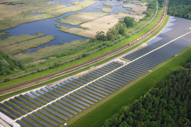 Luftaufnahme eines Solarparks mit Panelen, umgeben von Bäumen, Gras, Wasser und einer Bahn auf einem Gleis.