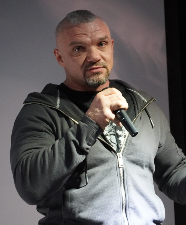 Komiker in einem schwarzen Anzug und einem weißen Hemd, der ein Mikrofon hält und selbstbewusst vor einer Wand steht.