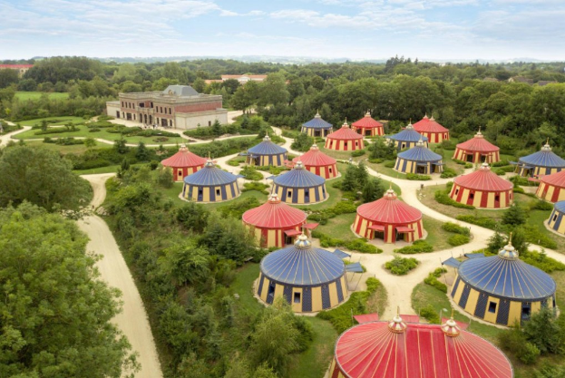 Luftaufnahme eines großen Zirkuszeltes auf einer grünen Wiese umgeben von Bäumen, Pflanzen und Gebäuden unter einem bewölkten Himmel.