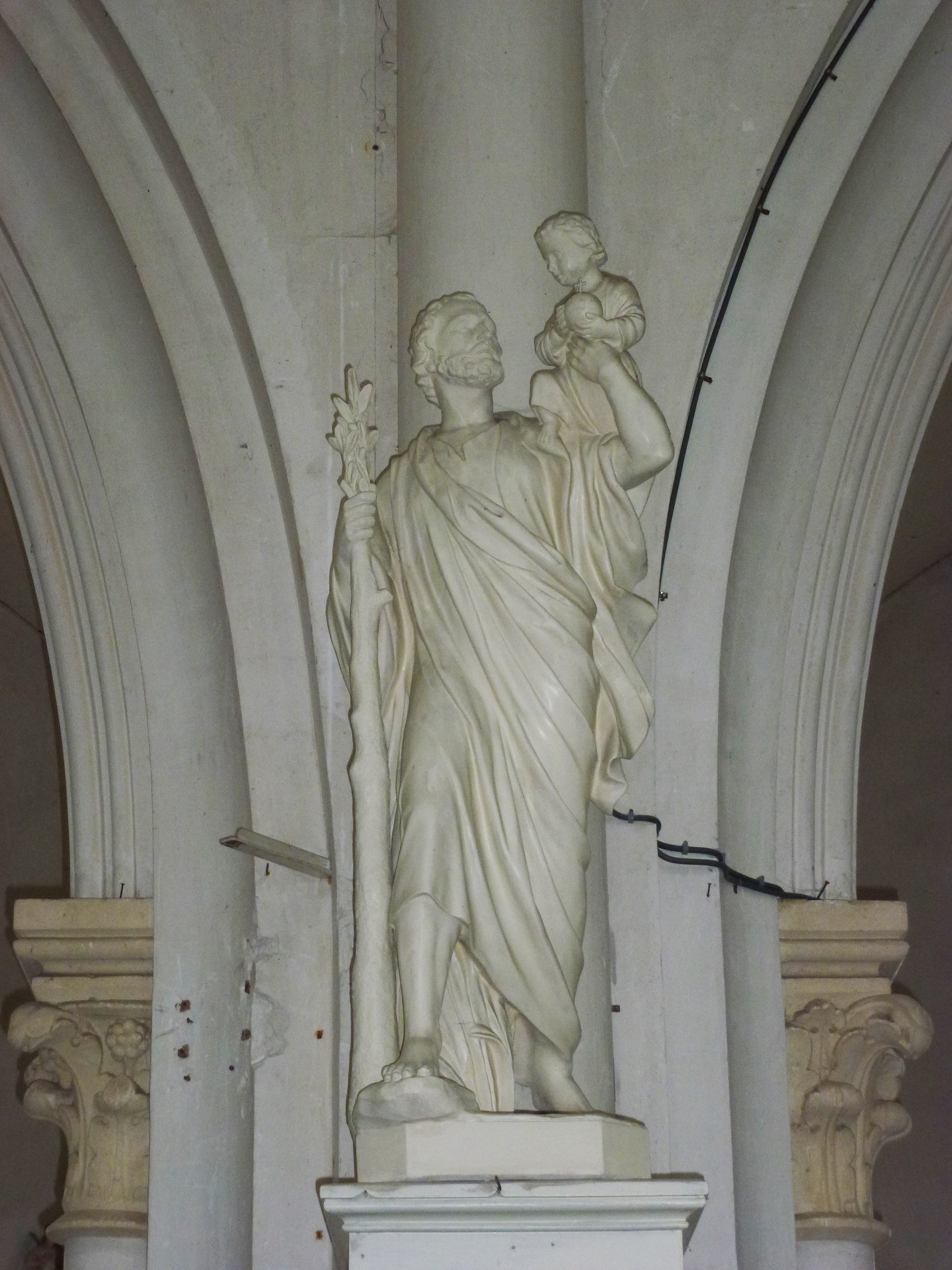 Statue eines gesegneten Mannes in einem langen Gewand, der ein Kind hält, das zu ihm aufschaut, mit zwei Säulen und einer Wand im Hintergrund, in einer Kirche.