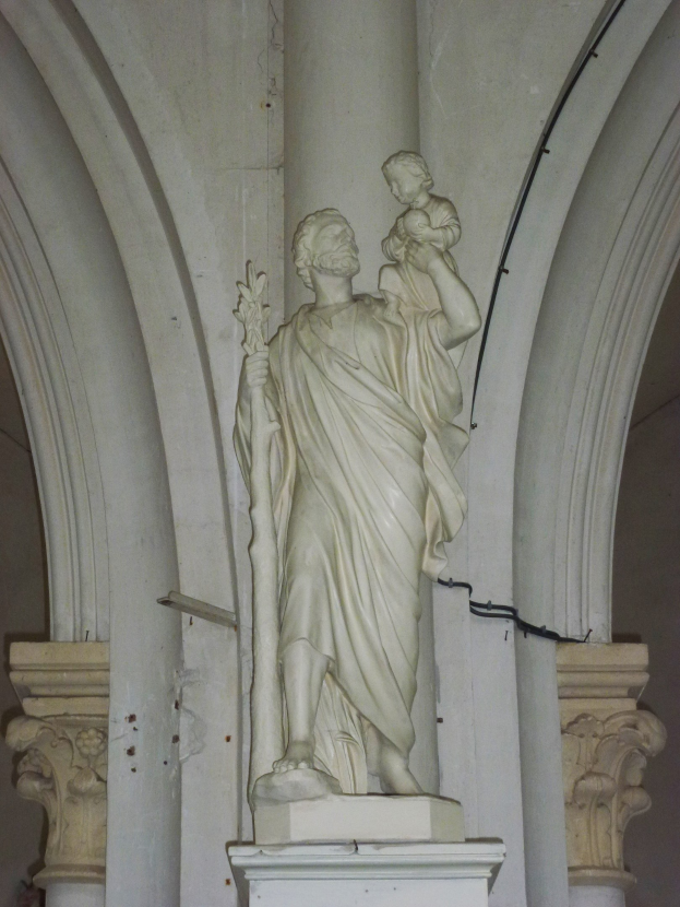 Statue eines gesegneten Mannes in einem langen Gewand, der ein Kind hält, das zu ihm aufschaut, mit zwei Säulen und einer Wand im Hintergrund, in einer Kirche.
