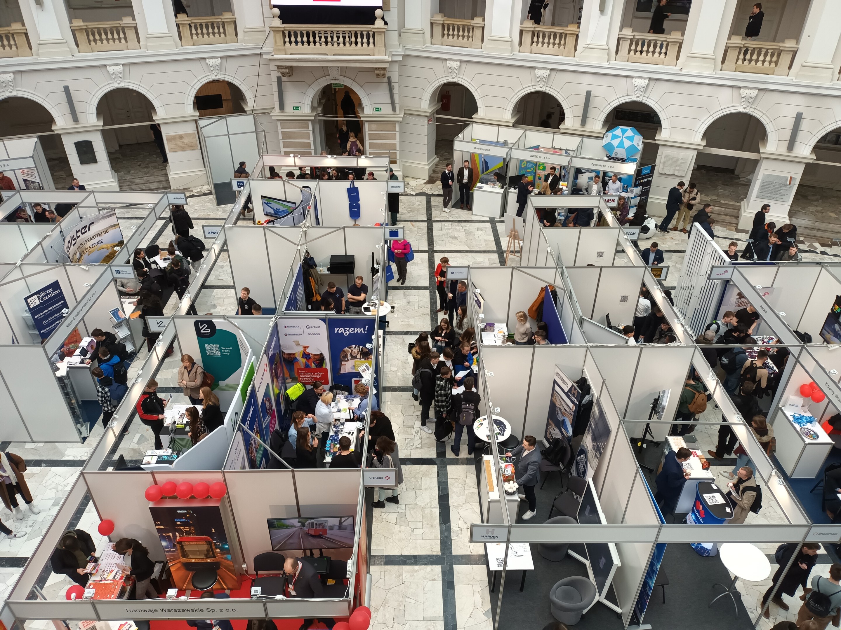 Eine große Gruppe von Menschen auf einer internationalen Fachmesse mit Ständen, Bannern, Tischen, Stühlen, Ballons und einem Gebäude mit Bögen, Säulen, Geländern und einem Hintergrundbild.
