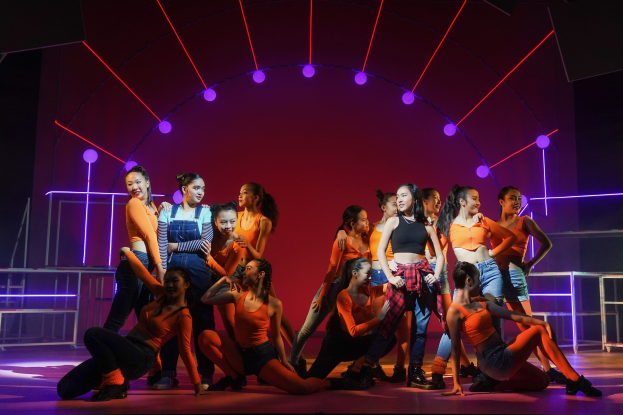 Eine Gruppe junger Tänzer in bunten Kostümen performs auf einer Bühne mit Geländern, die Arme erhoben, unter bunten Lichtern und geometrischen Bühnen-Dekorationen, während das Publikum applaudiert.