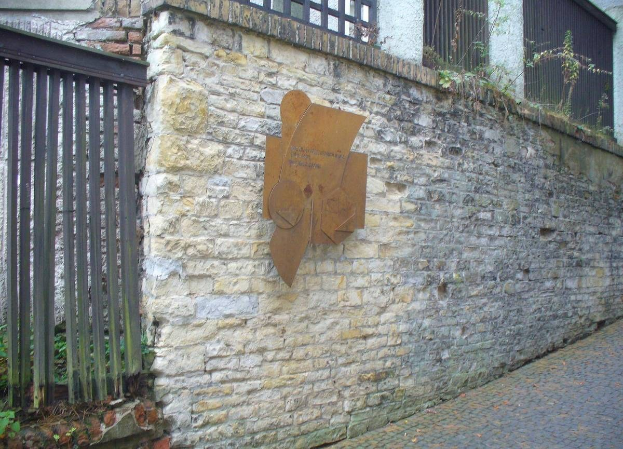 Steinmauer mit Metalltor auf der linken Seite, Gebäude mit Fenstern und Pflanzen im Hintergrund und eine Gedenktafel an der Wand zur Einweihung des alten Rathauses.