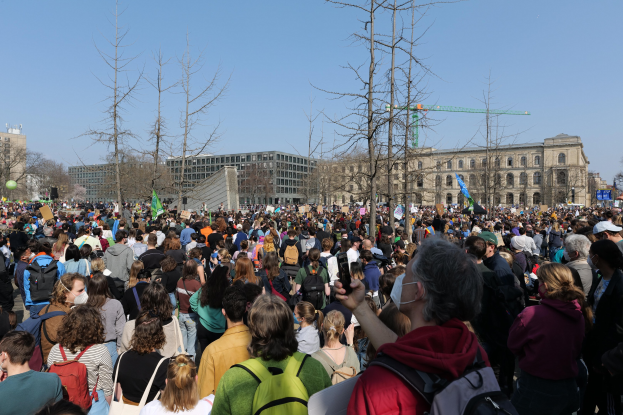 Eine große Menge Menschen steht vor einem Gebäude mit Fenstern, Bäumen und einem klaren Himmel, viele halten Schilder und tragen Taschen, was auf eine Klimawandel-Demonstration hindeutet.