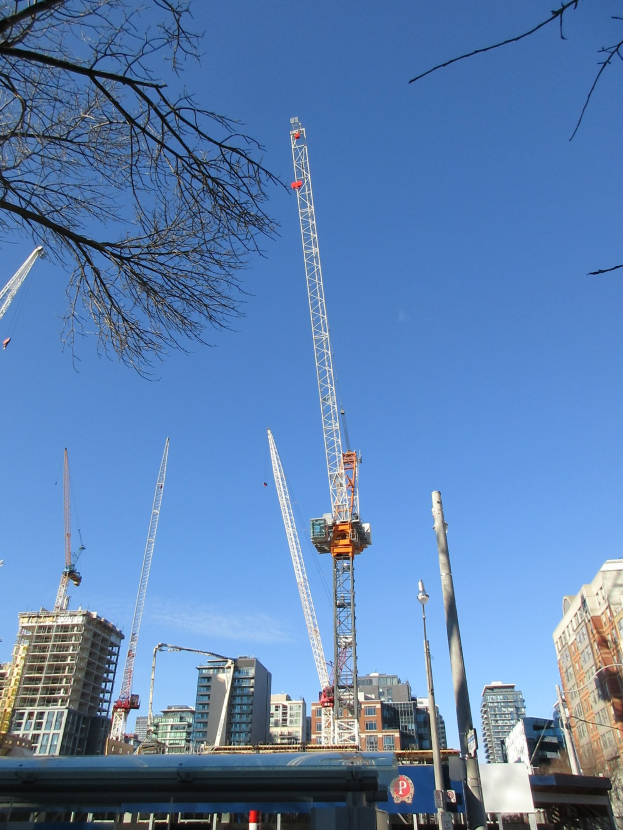 Baufahrzeuge, die über einer Stadtstraße aufragen, mit Gebäuden, Bäumen, Strommasten und Baustoffen im Vordergrund und einem sichtbaren Himmel.