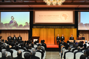 Eine Gruppe von Menschen in einem Raum mit einem Podium, Mikrofonen und einem großen Bildschirm, der Text und Bilder anzeigt, mit einer von einem Kronleuchter beleuchteten Decke darüber.