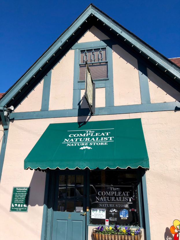 Ein Gebäude mit einem grünen Vordach, einer Tür, Fenstern, einem Schild mit der Aufschrift "Der Complete Naturalist Naturladen" und einem Korb mit blühenden Pflanzen unter einem bewölkten Himmel.