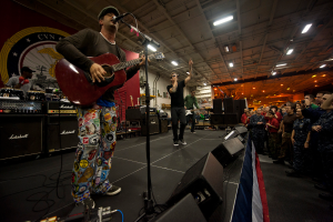 Chris Stapleton, mit einer Mütze bekleidet, spielt Gitarre auf der Bühne vor einem Publikum, umgeben von einem Mikrofon, Lautsprechern, einer Flagge und anderen Musikinstrumenten, mit einem Banner und Lichtern im Hintergrund.