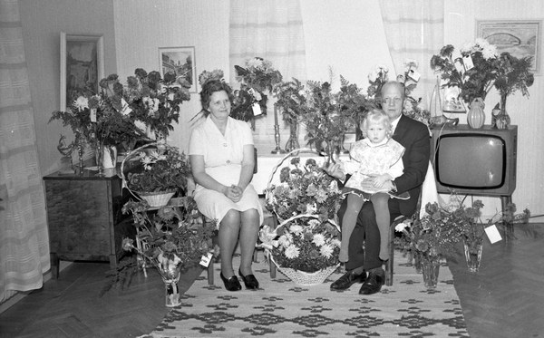 Schwarzes und weißes Foto einer Familie aus drei Personen - einem Mann, einer Frau und einem Kind - die auf einem Sofa vor einem Fernseher sitzen.