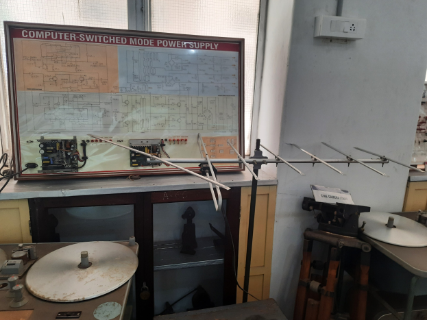 Ein unordentlicher Raum mit verschiedenen Elektronikgeräten, darunter eine etikettierte Leiterplatte auf einem Tisch, Metallstangen, eine wandmontierte Schalttafel, ein Fenster und Gegenstände in Schränken, die auf eine computerge.switchte Modus-Stromversorgung hinweisen.