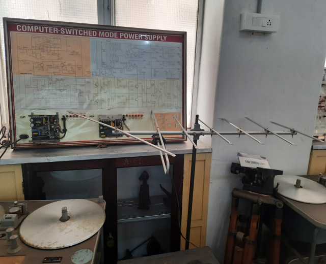 Ein unordentlicher Raum mit verschiedenen Elektronikgeräten, darunter eine etikettierte Leiterplatte auf einem Tisch, Metallstangen, eine wandmontierte Schalttafel, ein Fenster und Gegenstände in Schränken, die auf eine computerge.switchte Modus-Stromversorgung hinweisen.