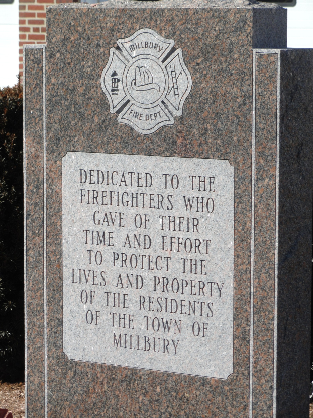 Denkmalstein mit einer Gedenktafel für Feuerwehrleute in Millbury, aufgestellt vor einem Backsteingebäude mit umgebender Vegetation.
