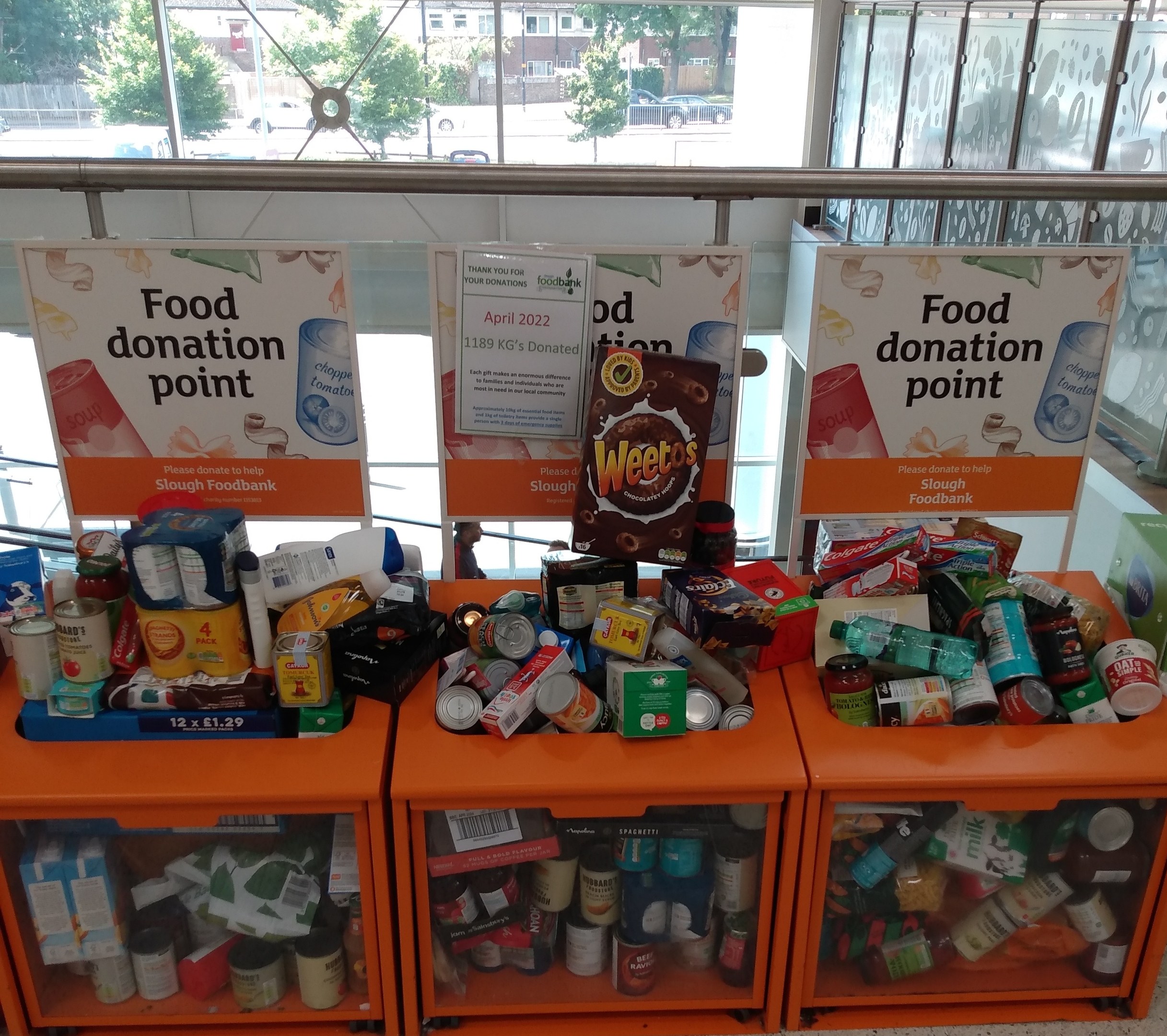 Orange Schübe mit Lebensmittelspenden gefüllt, darunter Flaschen, Dosen und Kisten, mit informativen Schildern im Hintergrund, vor einer Kulisse aus Fahrzeugen, Bäumen, Gebäuden und Himmel durch Glasfenster.