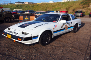 Ein Polizeiwagen auf einem Parkplatz mit anderen Autos, Gras, Pfählen, Drähten, Gebäuden und einem klaren blauen Himmel.