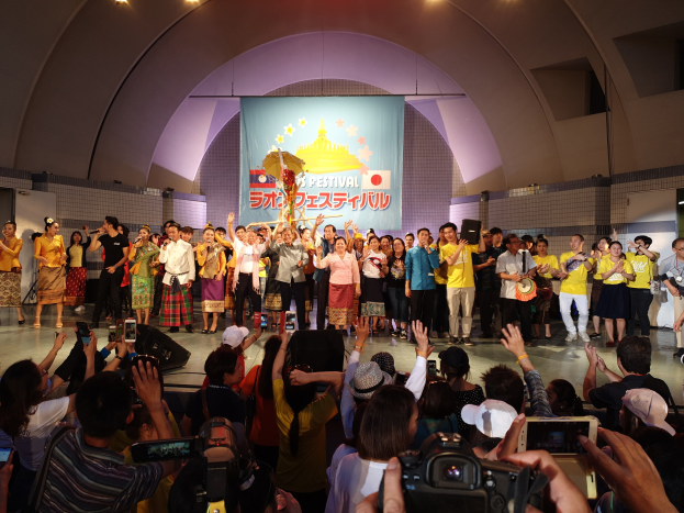 Eine Gruppe von Menschen auf einer Bühne mit Kameras, Handys und Musikinstrumenten während einer Festival-Eröffnung, mit Bannern, Wänden und Deckenlampen im Hintergrund.