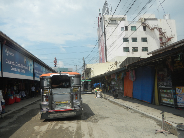 Ein Tuk-Tuk fährt auf einer Straße mit Geschäften auf beiden Seiten, im Hintergrund sind Gebäude, Strommasten mit Drähten und ein bewölkter Himmel zu sehen.