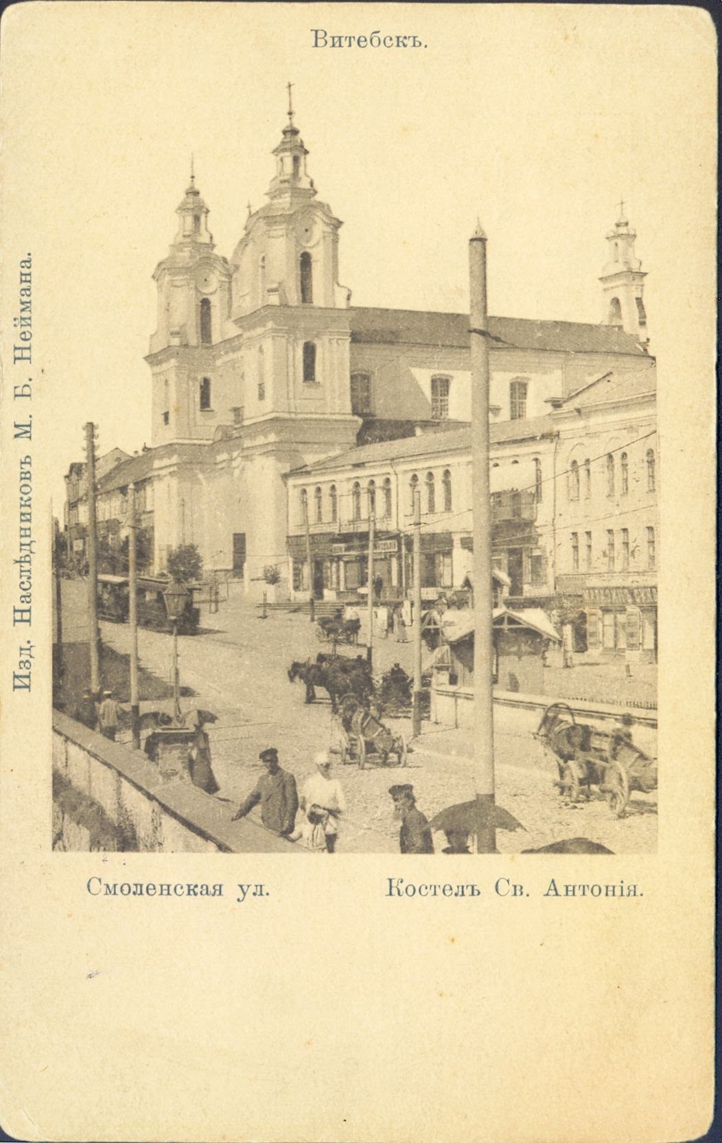 Altes Schwarz-Weiß-Foto einer Stadtstraße mit einer Kirche im Hintergrund, gesäumt von Bäumen und mit Gebäuden, Strommasten, Passanten, Pferdewagen und parkenden Pferdewagen auf der Straße, mit Text am unteren Rand.
