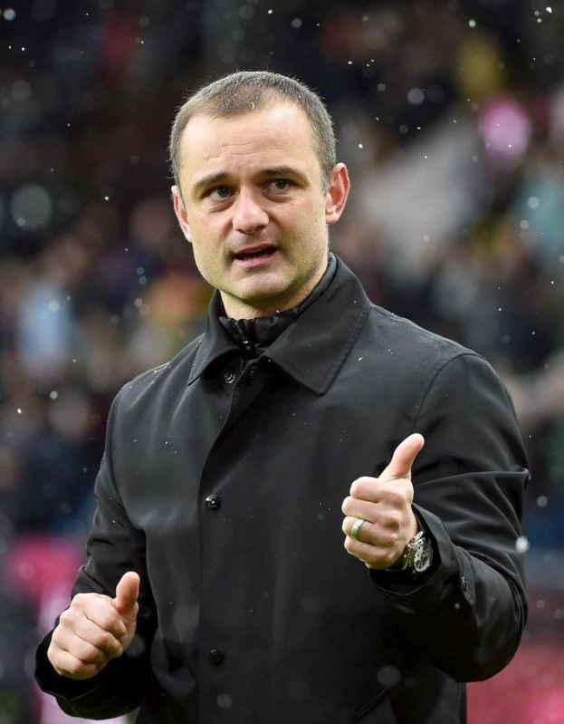 Ein Mann in einer schwarzen Jacke, der einen Daumen hoch hält, trägt eine Uhr und hat einen unscharfen Hintergrund.
