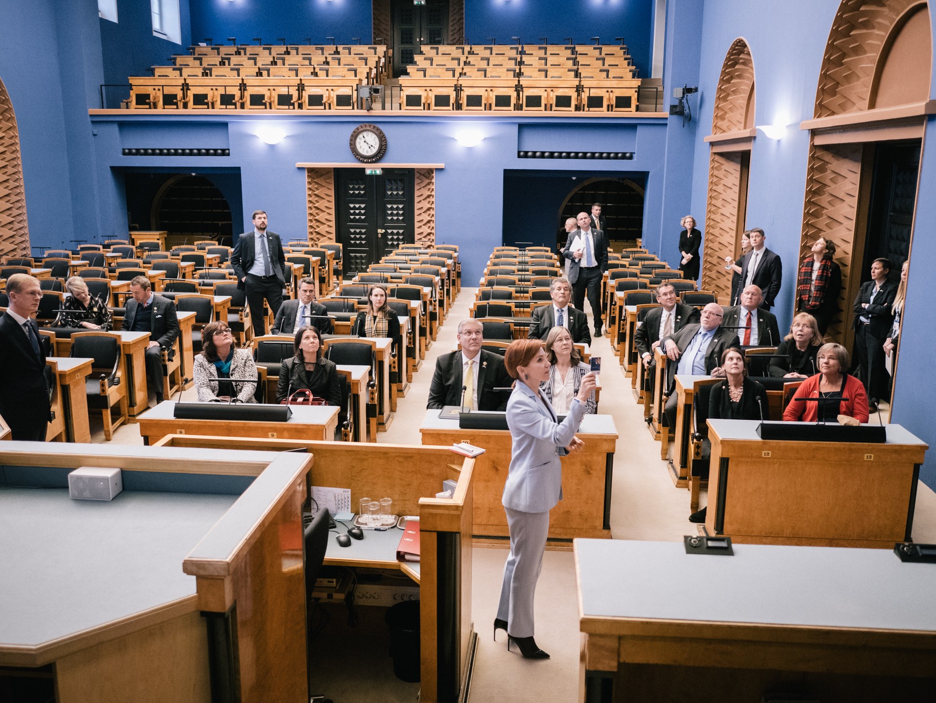 Frau hält Rede im schottischen Parlament, vor sitzenden und stehenden Zuhörern mit Mikrofonen, Gläsern, Papieren und anderen Gegenständen auf Tischen, mit einer Uhr und Deckenleuchten im Hintergrund.