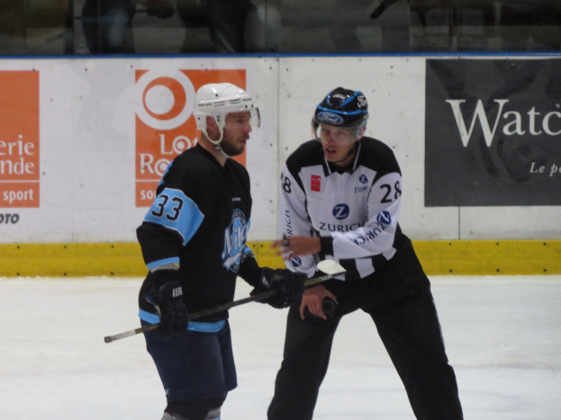 Zwei Männer mit Helmen und Eishockey-Ausrüstung spielen Eis hockey auf einem Eisplatz, mit Zuschauern durch die Glasscheibe und Text an der umliegenden Wand.