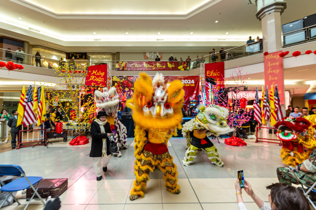 Vibrantes chinesisches Neujahrsfest in einem Einkaufszentrum mit Menschen in Kostümen, die tanzen, andere, die auf Stühlen sitzen und Handys verwenden, umgeben von Flaggen, Bannern, Dekorationsgegenständen, Geländern, Säulen und Deckenleuchten.