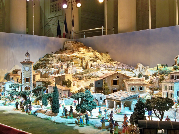 Ein detailliertes Miniatur-Weihnachtskrippen-Modell im Museum ausgestellt, mit Figuren, Gebäuden, Bäumen, Felsen, einem Glockenturm, Fahnen, Polen, Lichtern und einer Wand, beleuchtet von Museumsbeleuchtung.