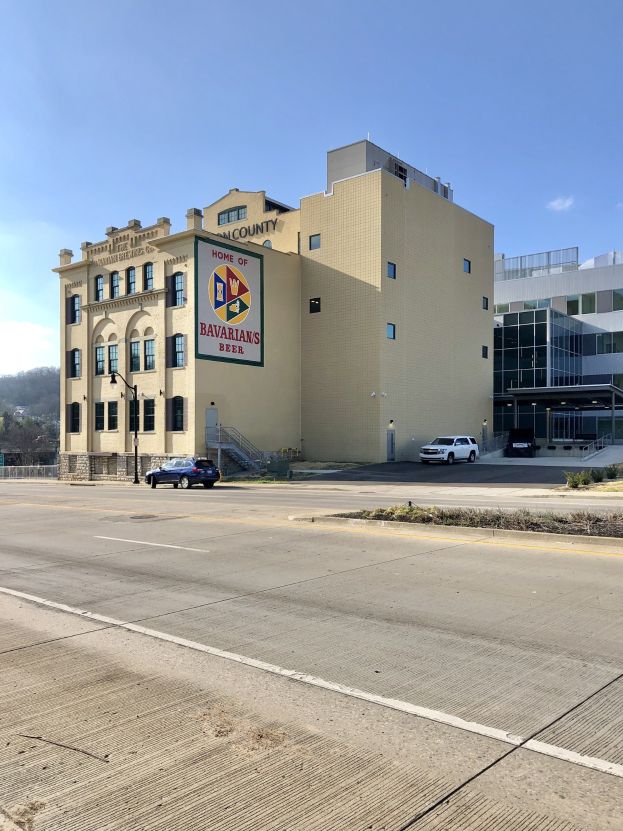 Großes Gebäude mit einer "Heimat der Bayern"-Aufschrift, umgeben von Fahrzeugen, Laternen, Pflanzen, Bäumen und einem bewölkten Himmel.