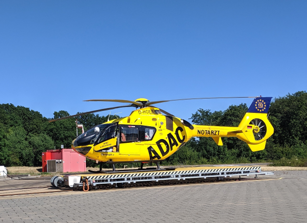 Ein gelber Hubschrauber auf einem Flughafen-Asphalt mit Bäumen und einem klaren blauen Himmel, umgeben von Gegenständen, die wahrscheinlich mit Luftrettungsdiensten in Deutschland in Verbindung stehen.