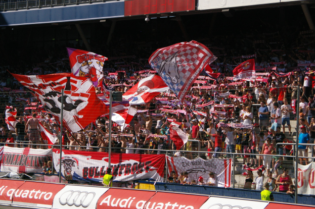 Eine große Menge in einem Stadion mit Flaggen, Transparenten, einem Metallzaun, einer Anzeigetafel, einem Bildschirm und Deckenleuchten.