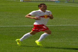 Ein Mann in weißem Hemd und roter Hose läuft auf einem Fußballfeld, im Hintergrund ist ein Tor zu sehen und ein paar Menschen stehen daneben, die Fußballschuhe tragen.